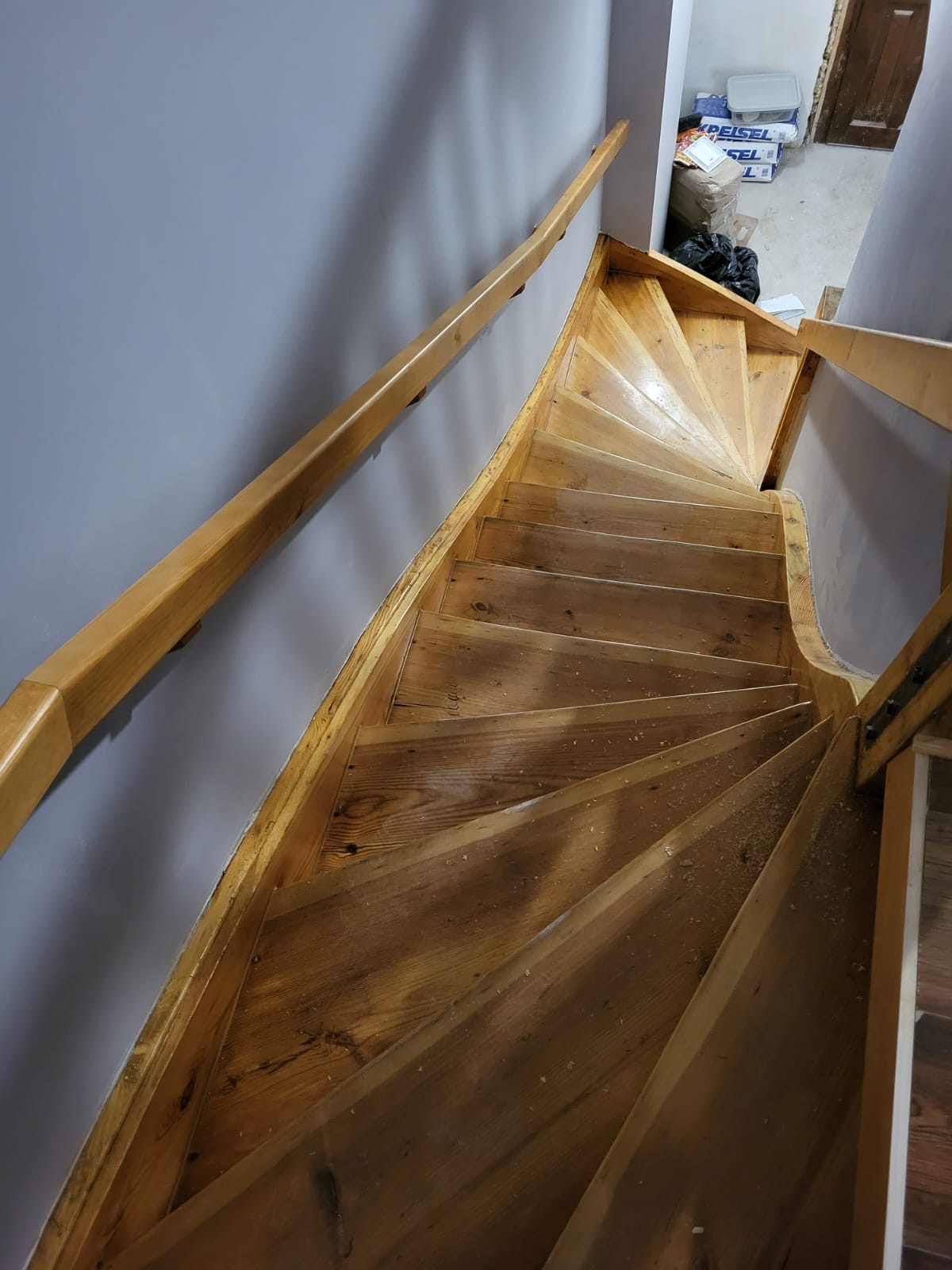 Drewniane schody kręcone podczas cyklinowania, widoczne ślady pyłu po obróbce, jasna barierka, szara ściana w tle.