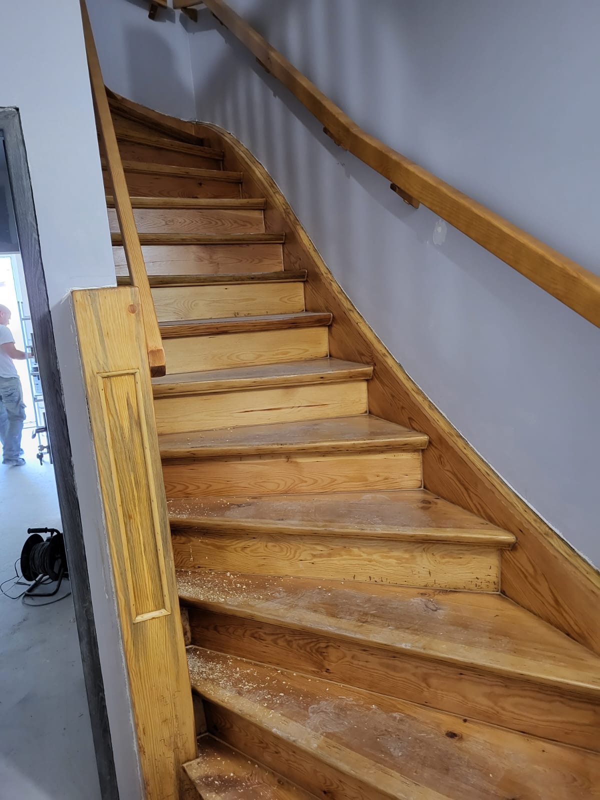 Drewniane schody z poręczą w trakcie renowacji, widoczne ślady szlifowania i pył, przygotowanie do kolejnych etapów prac wykończeniowych.