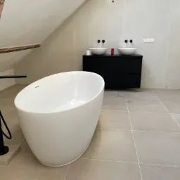 Nowoczesna łazienka na poddaszu z wolnostojącą białą wanną, czarną armaturą, dwiema umywalkami nablatowymi na czarnej szafce i beżowymi płytkami podłogowymi.