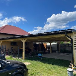 DrewLand - Konstrukcja z drewna w trakcie budowy, prawdopodobnie zadaszenie tarasu lub wiata garażowa, z widocznymi elementami szkieletu i częściowo zamontowaną ścianą z desek, na tle domu jednorodzinnego...