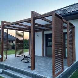 DrewLand - Drewniana pergola z lamelami w Ostrowcu Świętokrzyskim, z przezroczystym zadaszeniem, stojąca na szarym tarasie z kostki brukowej, widok na dom z ciemną dachówką.
