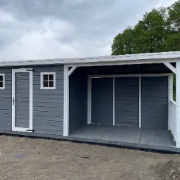 Szara konstrukcja z drewna z białymi detalami, zadaszona weranda z drewnianą podłogą, widok z zewnątrz na tle zieleni i pochmurnego nieba.