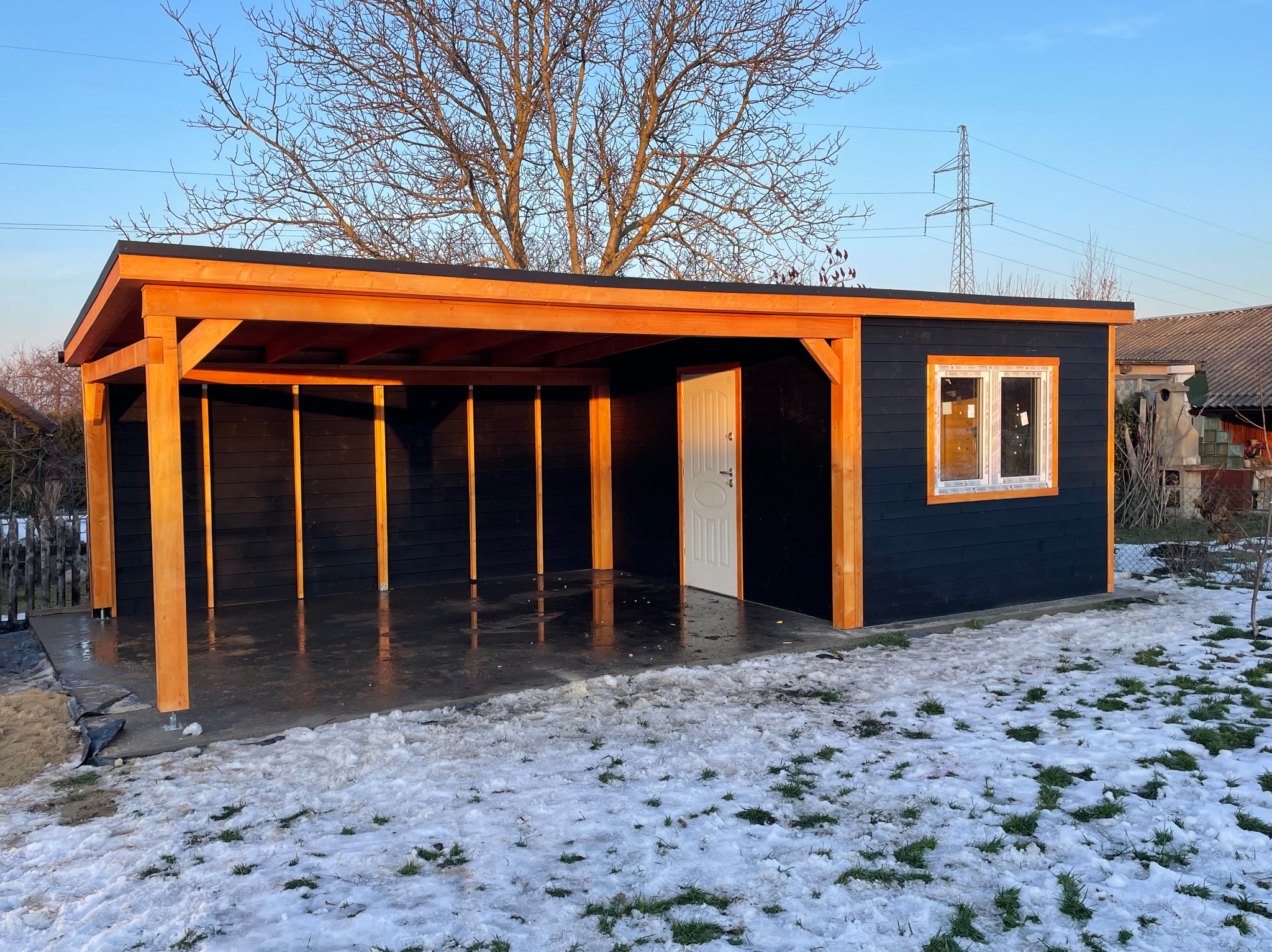 Ciemna, drewniana altana z jasnym drewnianym szkieletem i płaskim dachem, częściowo zadaszona, z białymi drzwiami i oknem, na betonowej wylewce, na tle zaśnieżonego trawnika i drzewa bez liści.