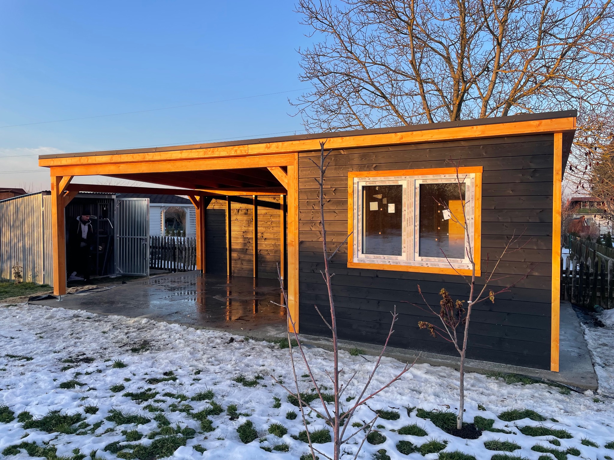 Ciemna, drewniana konstrukcja z jasnymi elementami wykończeniowymi, prawdopodobnie wiata garażowa lub mały budynek gospodarczy, częściowo pokryta śniegiem, z widoczną osobą w środku
