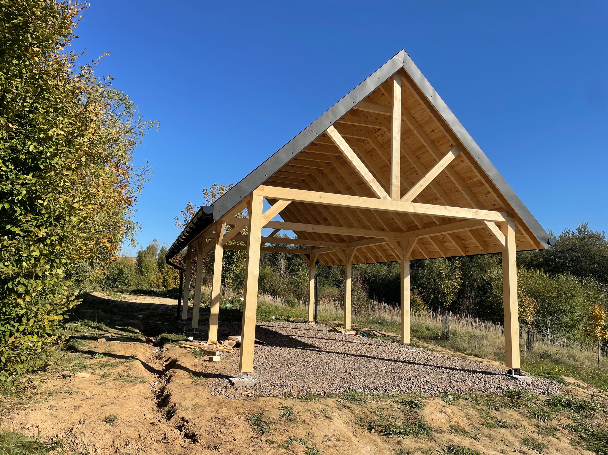 Konstrukcja z drewna w trakcie budowy, z widocznym szkieletem dachu i kolumnami, posadowiona na żwirowym podłożu, z otaczającą zielenią i błękitnym niebem w tle.
