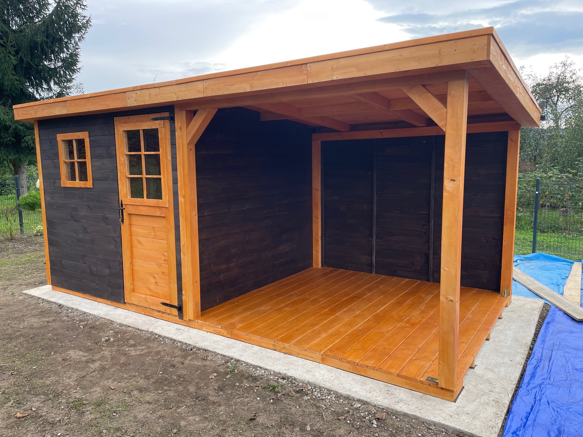 Nowoczesna drewniana konstrukcja z zadaszeniem, pomalowana na ciemny brąz z jasnymi elementami wykończeniowymi, ustawiona na betonowej wylewce. Widoczne drzwi z szybkami i małe okno.