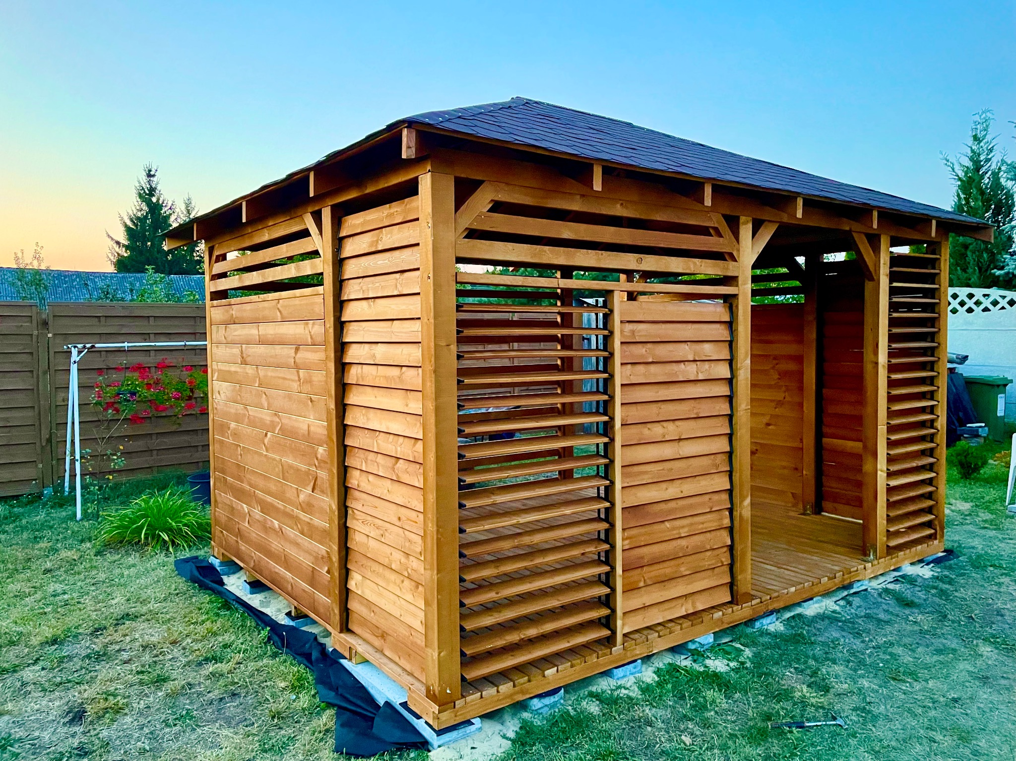 Drewniana konstrukcja z zadaszeniem na trawie, z elementami lamelowymi, prawdopodobnie w trakcie montażu, widoczne zabezpieczenia podłoża.