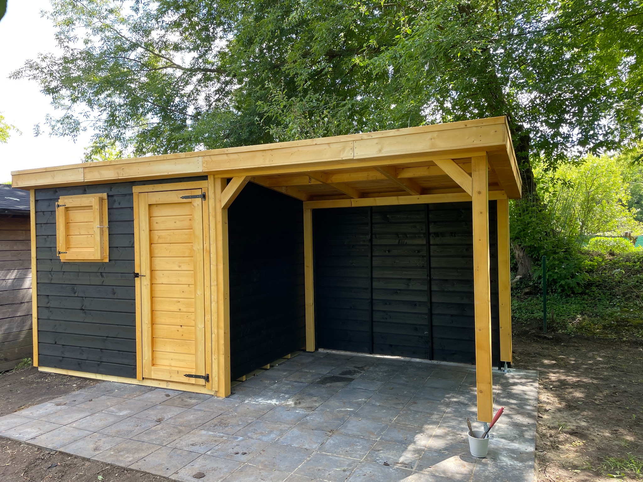 Czarno-żółta konstrukcja z drewna na zewnątrz, z częścią zamykaną drzwiami i oknem, a częścią otwartą, stojąca na szarych płytkach chodnikowych, z drzewami w tle i wiadrem z pędzlami obok.