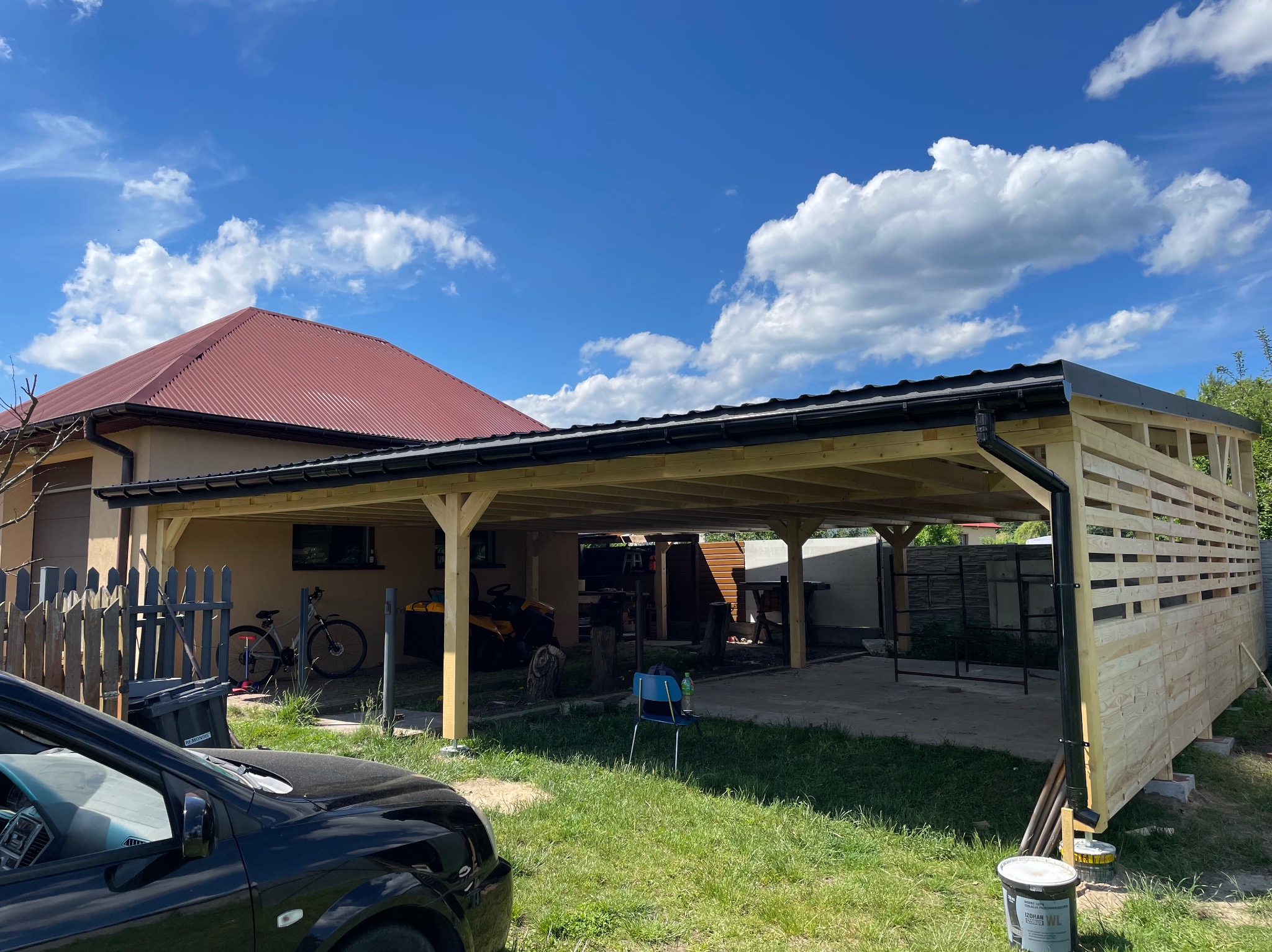 Konstrukcja z drewna w trakcie budowy, prawdopodobnie zadaszenie tarasu lub wiata garażowa, z widocznymi elementami szkieletu i częściowo zamontowaną ścianą z desek, na tle domu jednorodzinnego...