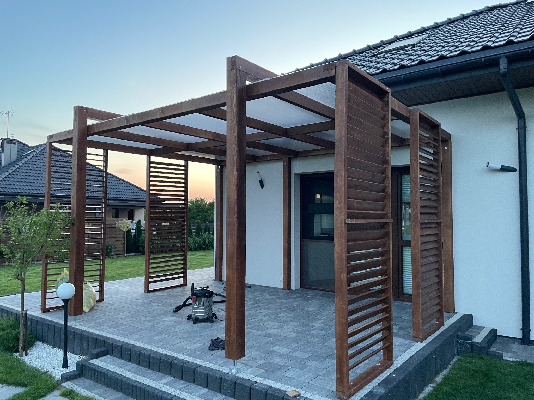 Drewniana pergola z lamelami w Ostrowcu Świętokrzyskim, z przezroczystym zadaszeniem, stojąca na szarym tarasie z kostki brukowej, widok na dom z ciemną dachówką.