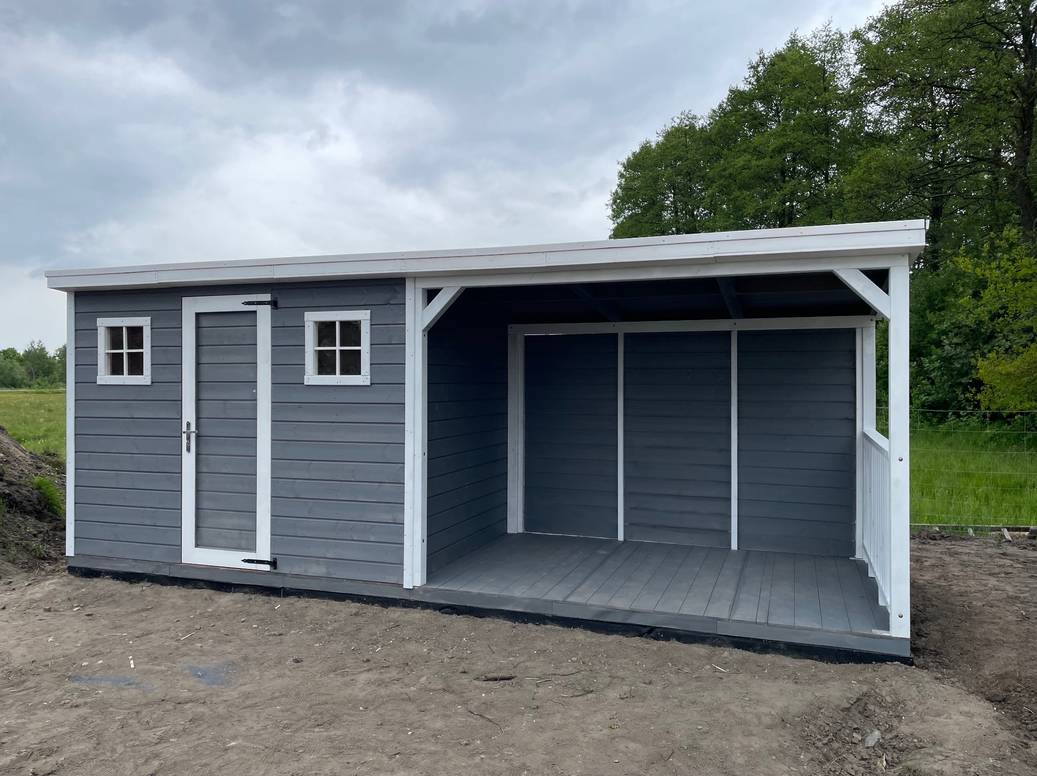 Szara konstrukcja z drewna z białymi detalami, zadaszona weranda z drewnianą podłogą, widok z zewnątrz na tle zieleni i pochmurnego nieba.