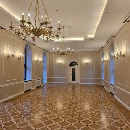 RS BUD - Eleganckie wnętrze sali balowej z parkietem w szachownicę, kremowymi ścianami z ozdobnymi listwami, złotymi żyrandolami i kinkietami, oraz wysokimi, łukowatymi oknami.