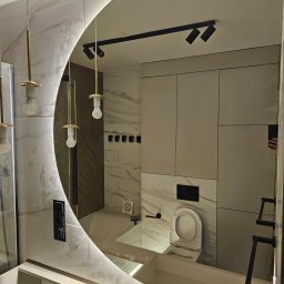 RS BUD - Nowoczesna łazienka z okrągłym lustrem podświetlanym LED, widoczne marmurowe płytki, minimalistyczna toaleta i oświetlenie punktowe na suficie.