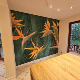 RS BUD - Fototapeta z motywem rajskich ptaków na ciemnozielonym tle z liśćmi, w jasnym wnętrzu z drewnianym stołem i widokiem na ogród przez okno.