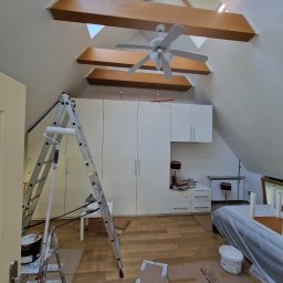 RS BUD - Poddasze w trakcie malowania: drabina z wałkiem malarskim oparta o ścianę, zabezpieczona podłoga kartonem, widoczne drewniane belki stropowe i okna dachowe.