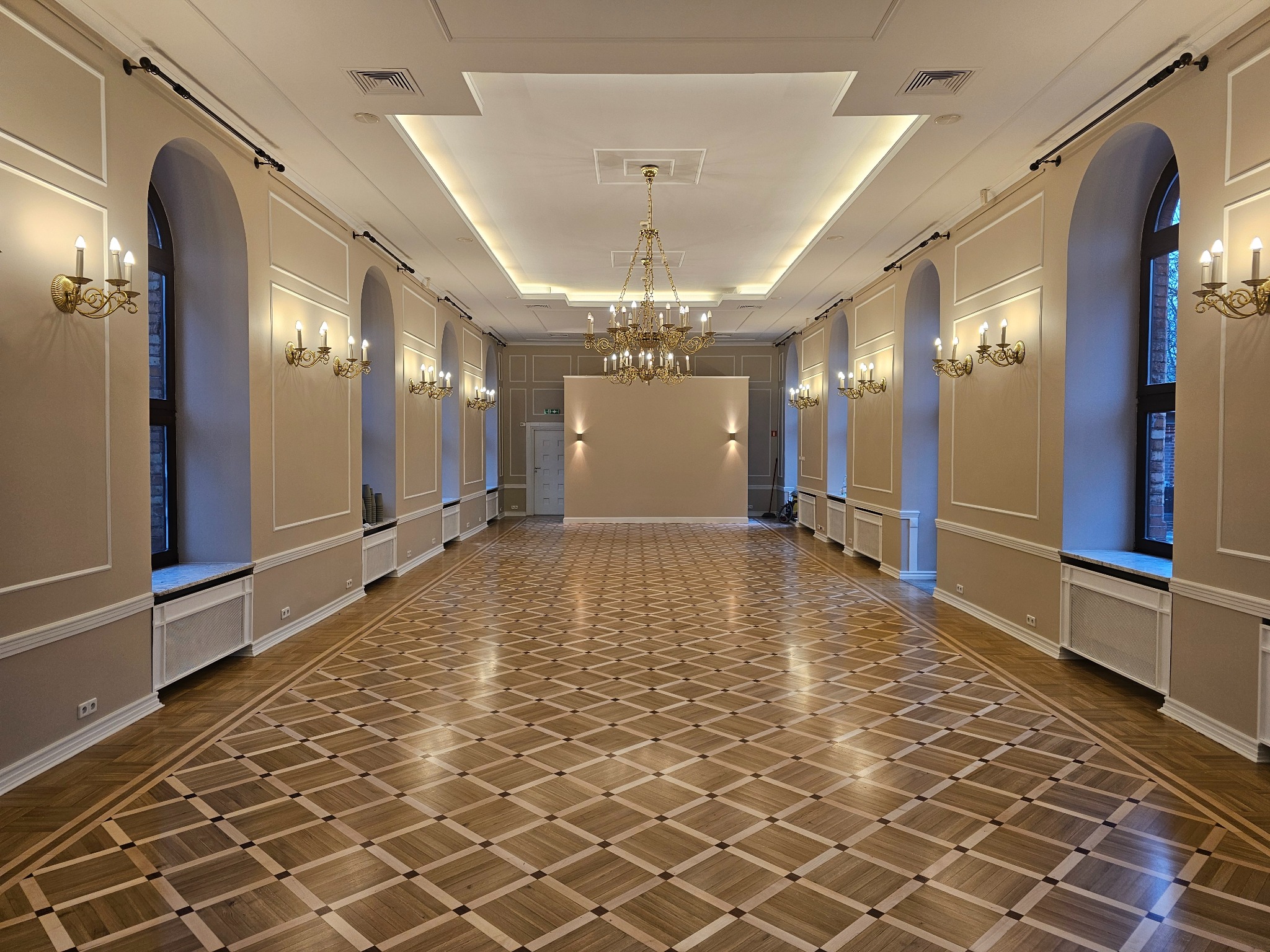 Przestronne wnętrze z parkietem w geometryczny wzór, kremowymi ścianami z dekoracyjnymi listwami, żyrandolem i kinkietami w stylu retro.