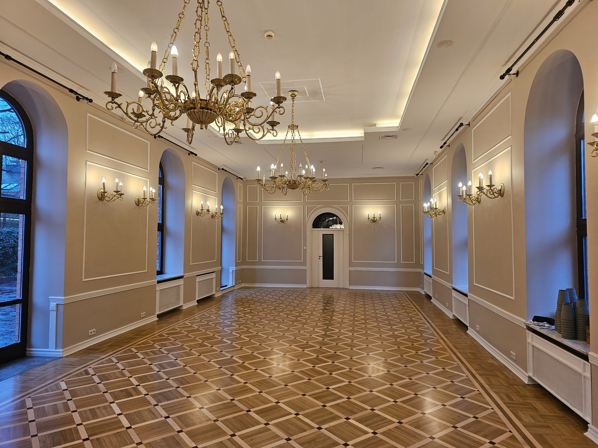 Eleganckie wnętrze sali balowej z parkietem w szachownicę, kremowymi ścianami z ozdobnymi listwami, złotymi żyrandolami i kinkietami, oraz wysokimi, łukowatymi oknami.