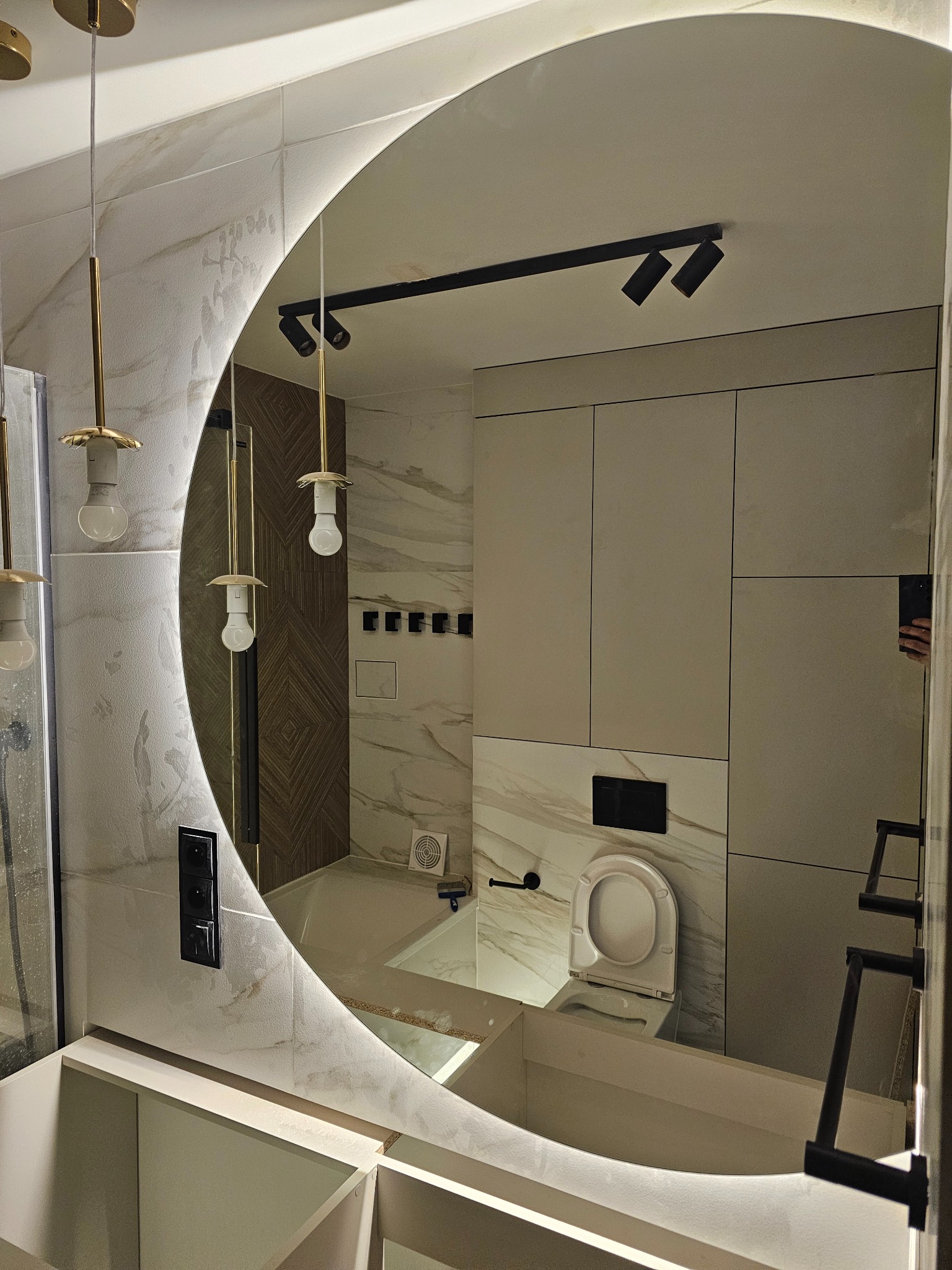 Nowoczesna łazienka z okrągłym lustrem podświetlanym LED, widoczna armatura w kolorze czarnym, płytki imitujące marmur i dekoracyjna ściana z drewnianymi panelami.