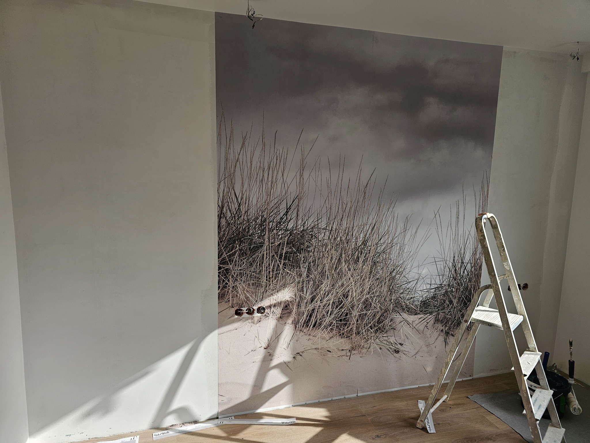 Ściana w trakcie remontu z fragmentem fototapety przedstawiającej wydmę z trawami, widoczne gniazdka elektryczne i drabina malarska