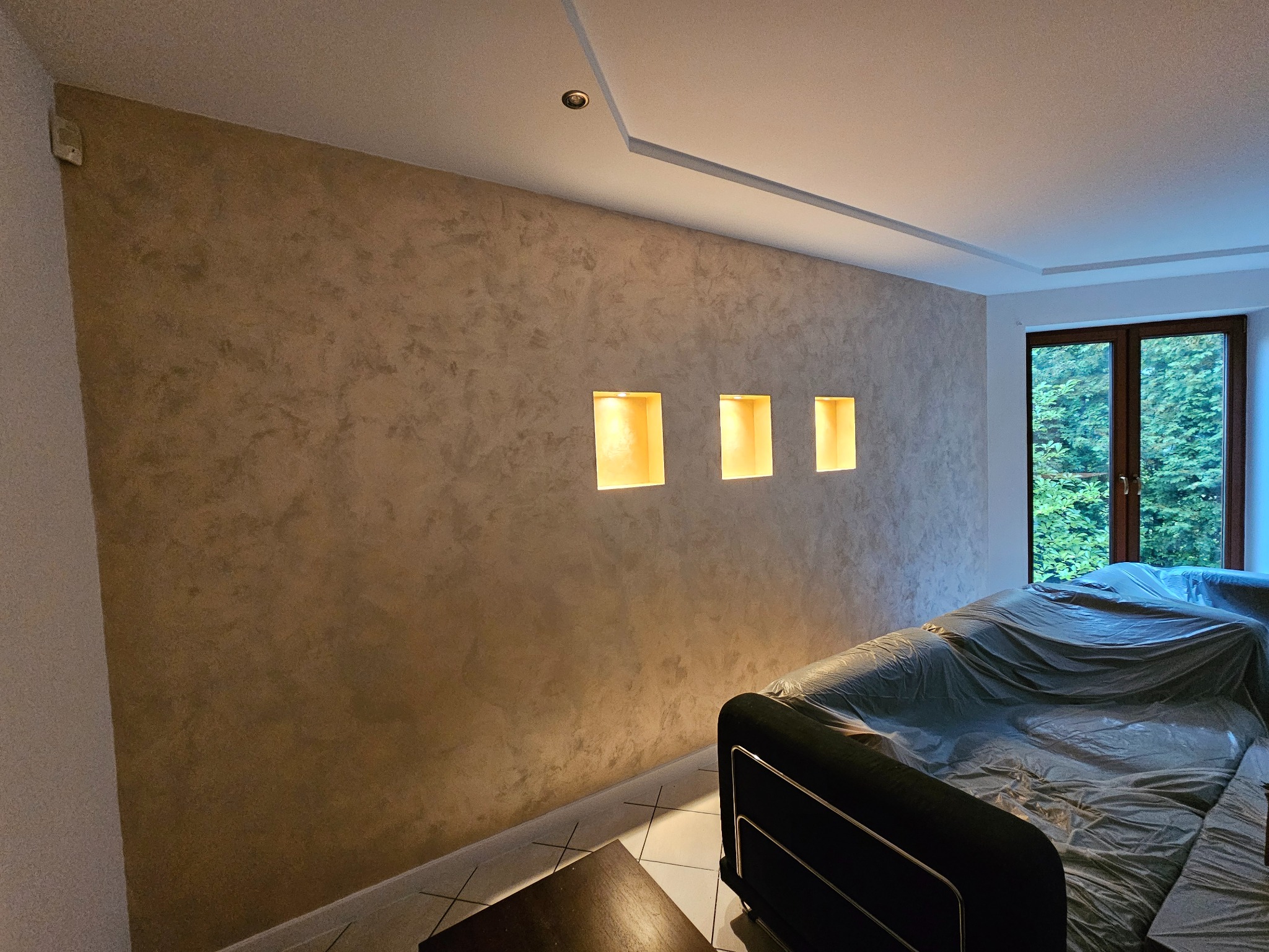 Ściana w pokoju po nałożeniu dekoracyjnej, beżowo-brązowej gładzi z wnękami oświetlonymi ciepłym światłem, meble przykryte folią ochronną.