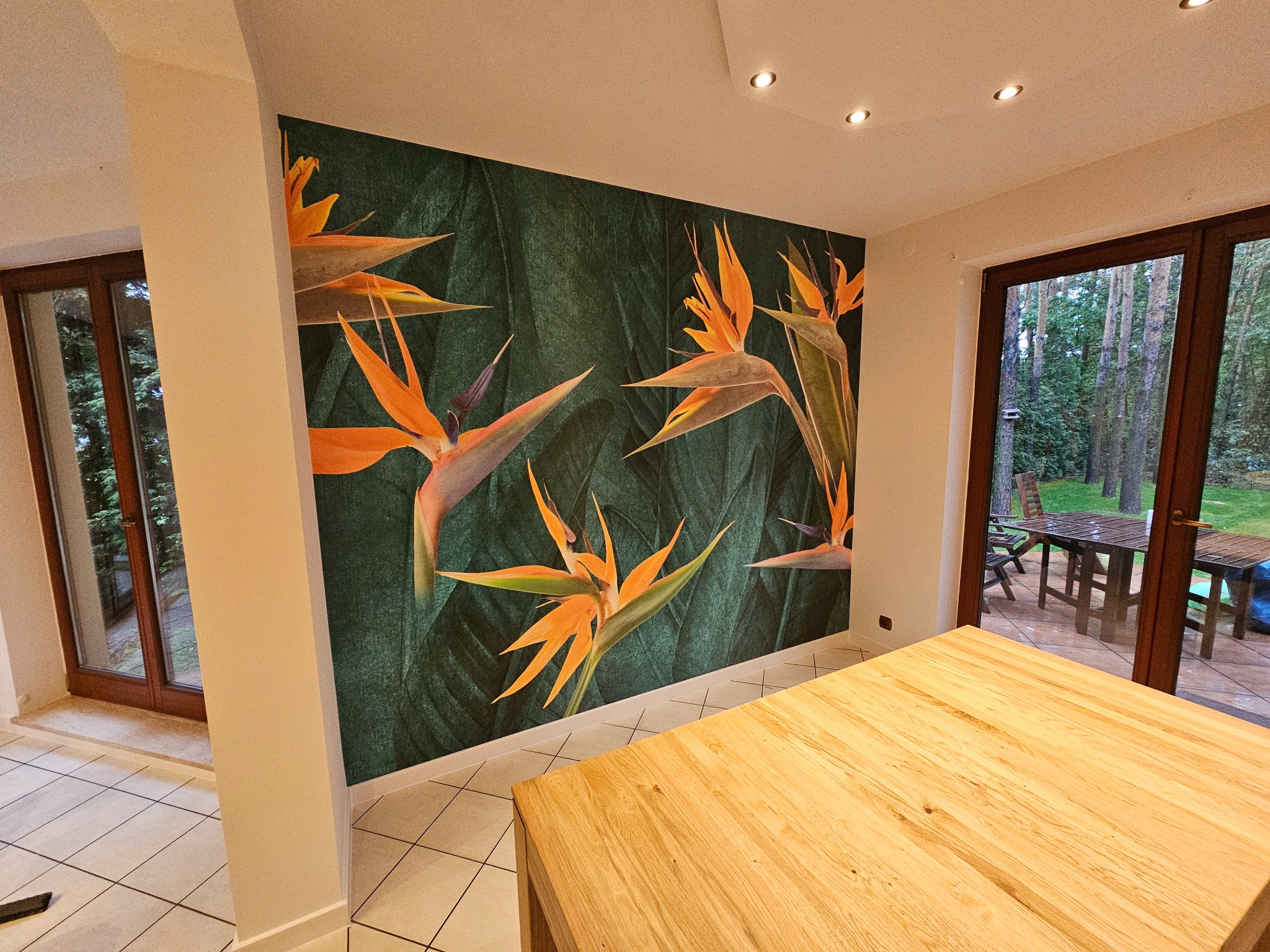 Fototapeta z motywem rajskich ptaków na ciemnozielonym tle z liśćmi, w jasnym wnętrzu z drewnianym stołem i widokiem na ogród przez okno.