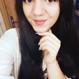 Portret uśmiechniętej brunetki z brązowymi oczami, w białej koszuli, z dłonią pod brodą i szarymi paznokciami.