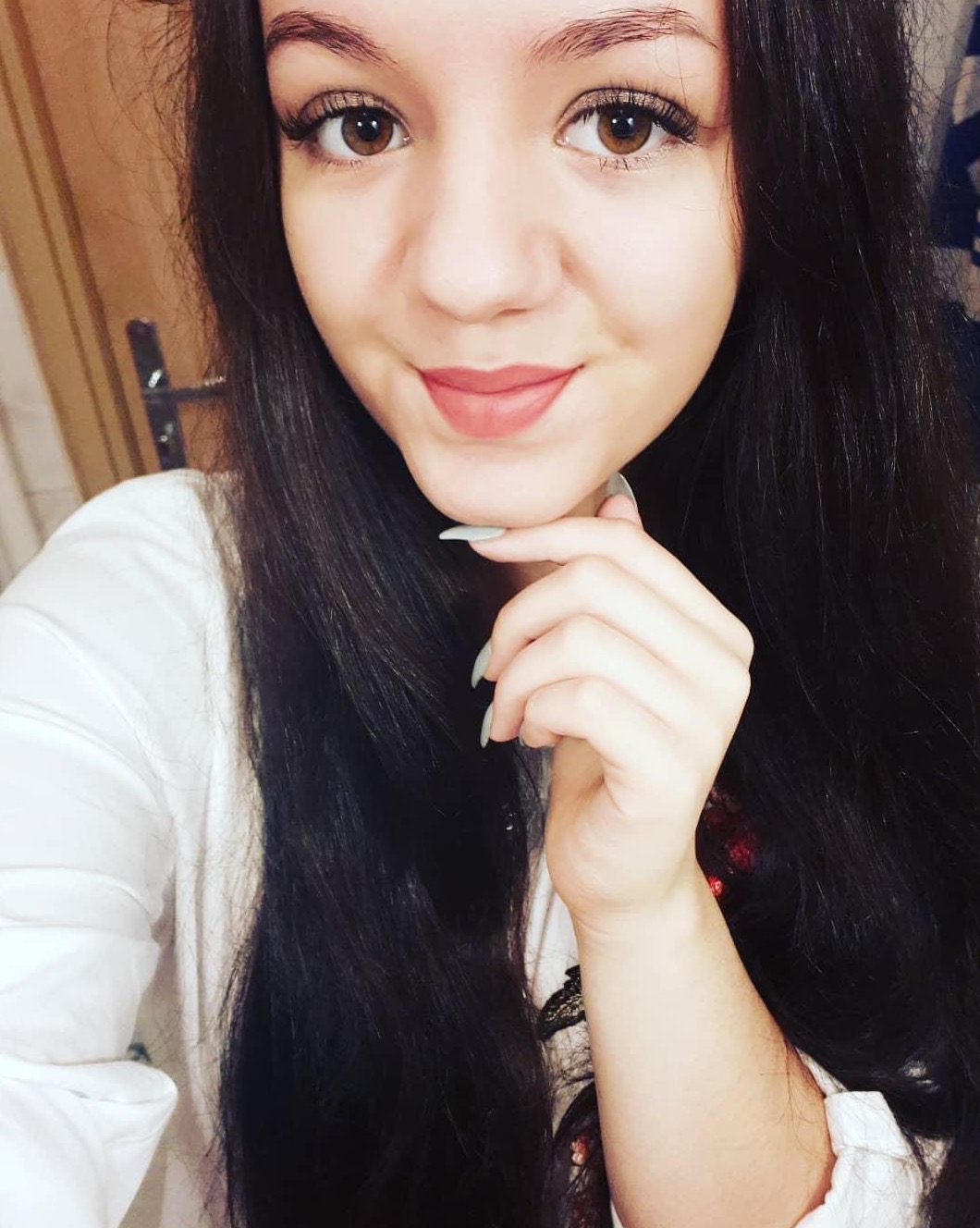 Portret uśmiechniętej brunetki z brązowymi oczami, w białej koszuli, z dłonią pod brodą i szarymi paznokciami.