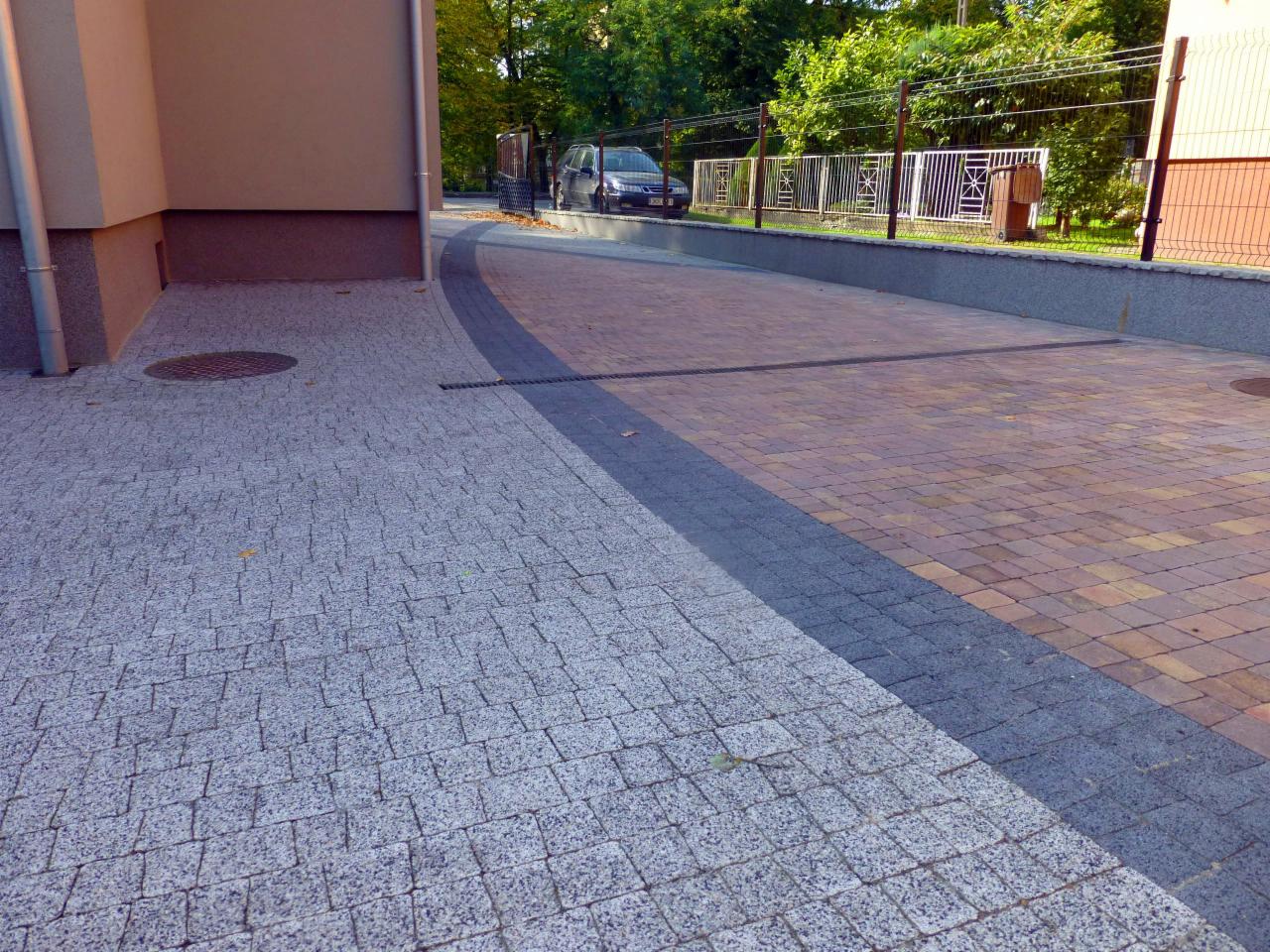 Ułożona kostka brukowa w dwóch kolorach, szarym i ceglastym, oddzielona ciemniejszym pasem. Na dalszym planie budynek i zaparkowany samochód.