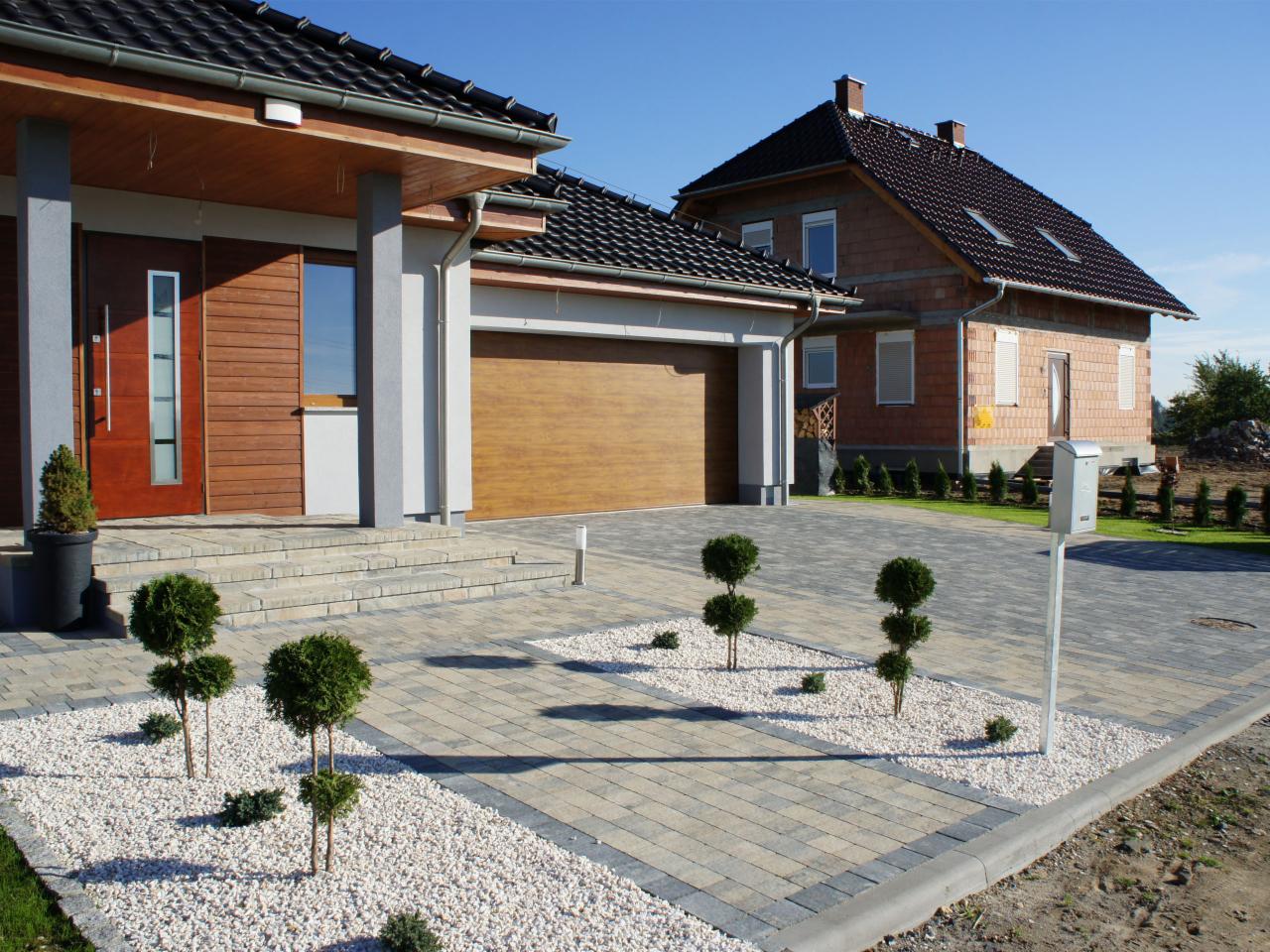 Nowoczesna kostka brukowa w jasnych odcieniach, zdobiąca podjazd do domu z drewnianą bramą garażową. Małe drzewka i biały żwir dodają elegancji.