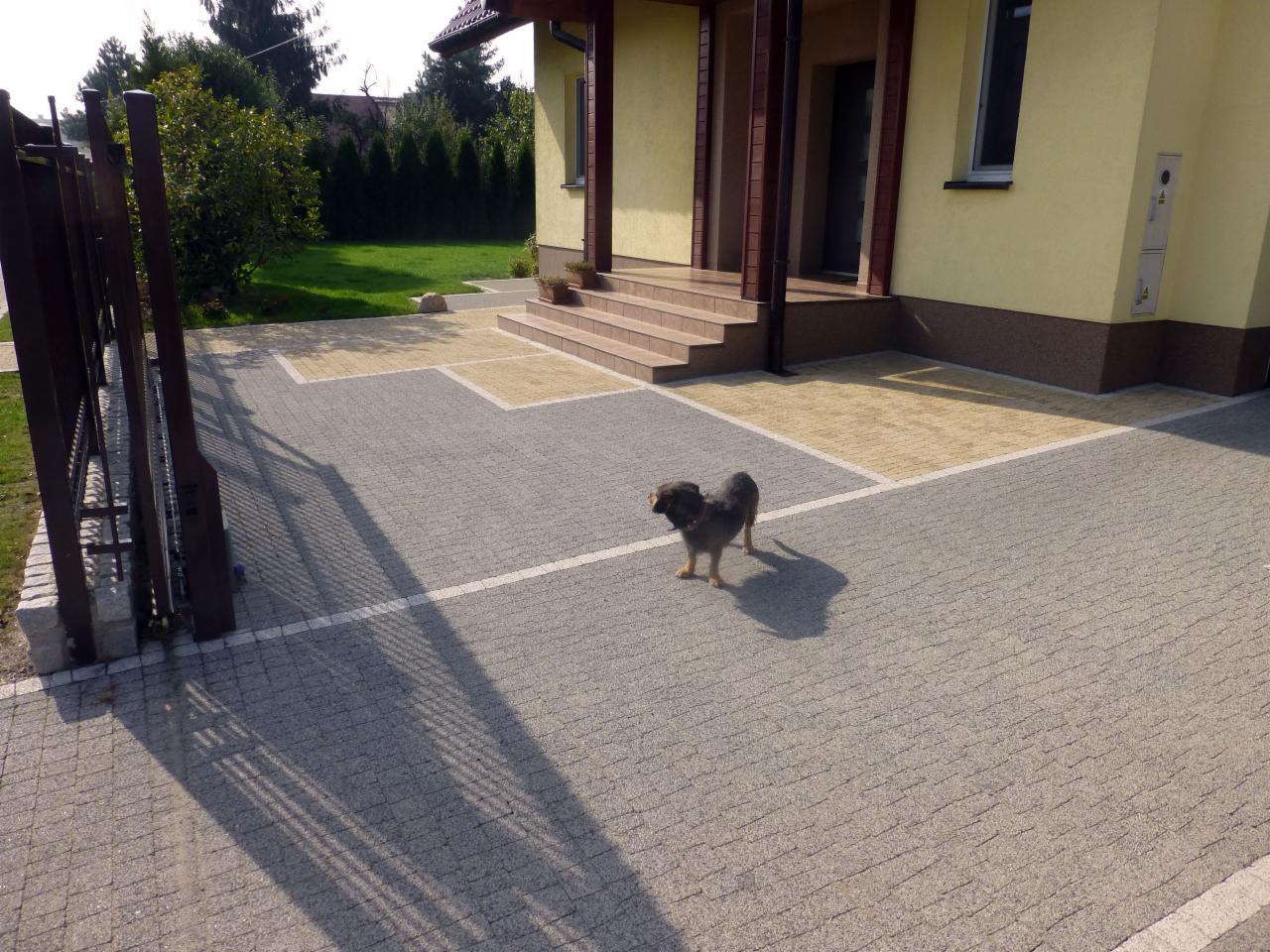 Ułożona kostka brukowa w dwóch odcieniach szarości i beżu tworząca wzór przed domem. W tle widoczny dom z beżową elewacją i brązowymi elementami wykończeniowymi.