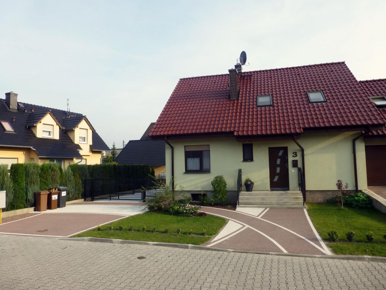 Dom jednorodzinny z czerwonym dachem i starannie zagospodarowanym terenem wokół. Widoczny chodnik z kostki brukowej i zadbany trawnik z roślinami ozdobnymi.
