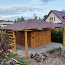 Drewniana konstrukcja z zadaszeniem na betonowej wylewce, widok z zewnątrz na tle domu jednorodzinnego i zachmurzonego nieba. Na pierwszym planie fragment ogrodu z roślinnością i stertą drewna.
