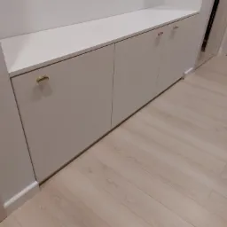 Biała zabudowa meblowa z trzema szafkami z owalnymi, złotymi uchwytami, umieszczona w przedpokoju z panelami podłogowymi w odcieniach beżu.