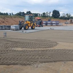 Taras Kutsenko - Żółta ładowarka obok palet z kostką brukową i pracowników w odblaskowych ubraniach na placu budowy z częściowo ułożoną kostką, widoczne ślady opon na piasku.