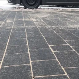 Ułożona, szara kostka brukowa z widocznymi szczelinami wypełnionymi piaskiem, z fragmentami zaparkowanych samochodów w tle.