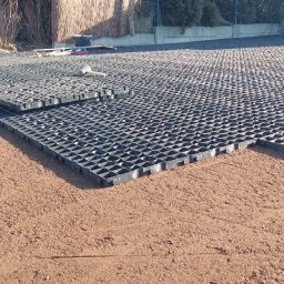 Taras Kutsenko - Częściowo ułożona, czarna, plastikowa kratka trawnikowa na podsypce z czerwonego piasku; w tle widoczny pracownik i narzędzia.