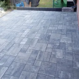 Ułożona szara kostka brukowa na posesji, widok z góry. Różne rozmiary kostek tworzą wzór. W tle siatka ogrodzeniowa i zieleń. Krawężnik z ciemnej kostki.