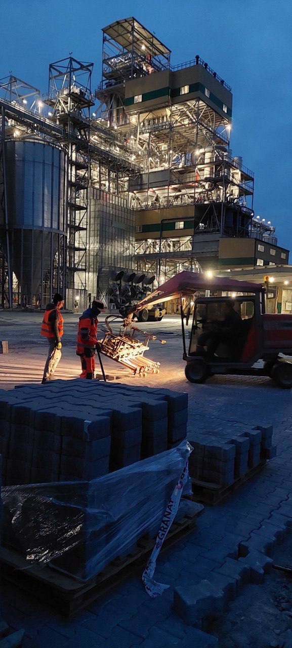 Dwóch pracowników w pomarańczowych kamizelkach układa kostkę brukową za pomocą specjalistycznej maszyny na tle oświetlonej hali przemysłowej o zmierzchu. Na pierwszym planie paleta z kostką brukową...