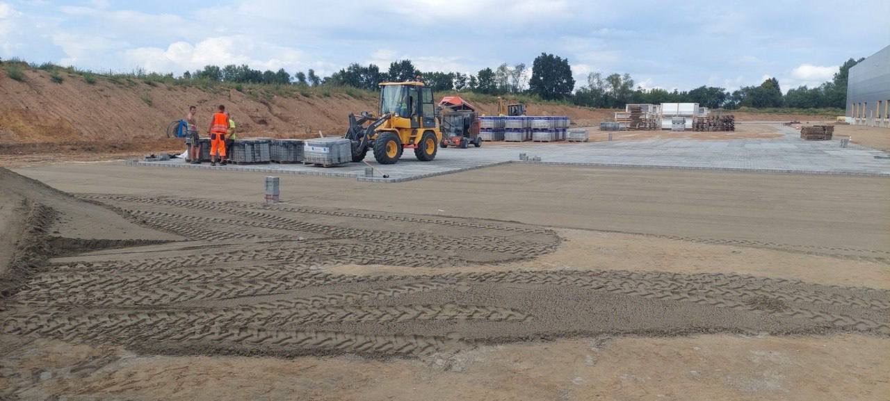 Żółta ładowarka obok palet z kostką brukową i pracowników w odblaskowych ubraniach na placu budowy z częściowo ułożoną kostką, widoczne ślady opon na piasku.