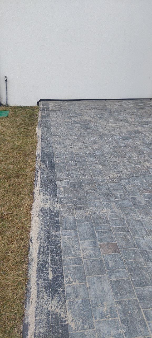 Świeżo ułożona, szara kostka brukowa z ciemniejszym obramowaniem, częściowo zasypana piaskiem, przylegająca do trawnika i białej ściany budynku.