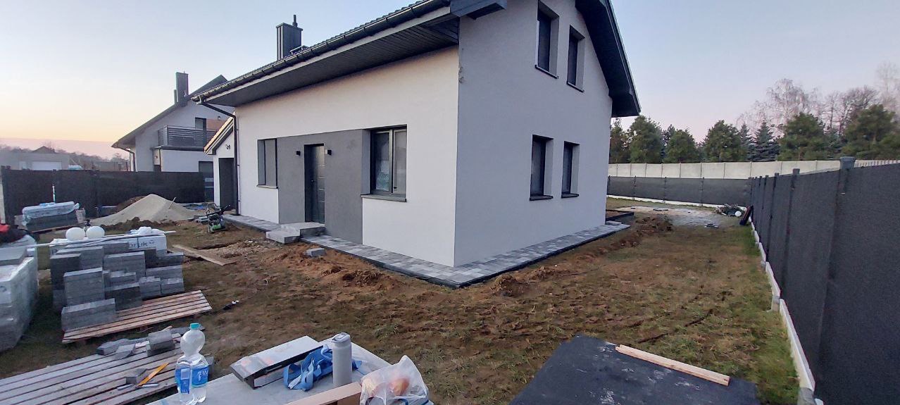 Nowoczesny dom jednorodzinny z szarą kostką brukową wokół, widoczne materiały budowlane i narzędzia na pierwszym planie, szare ogrodzenie w tle.