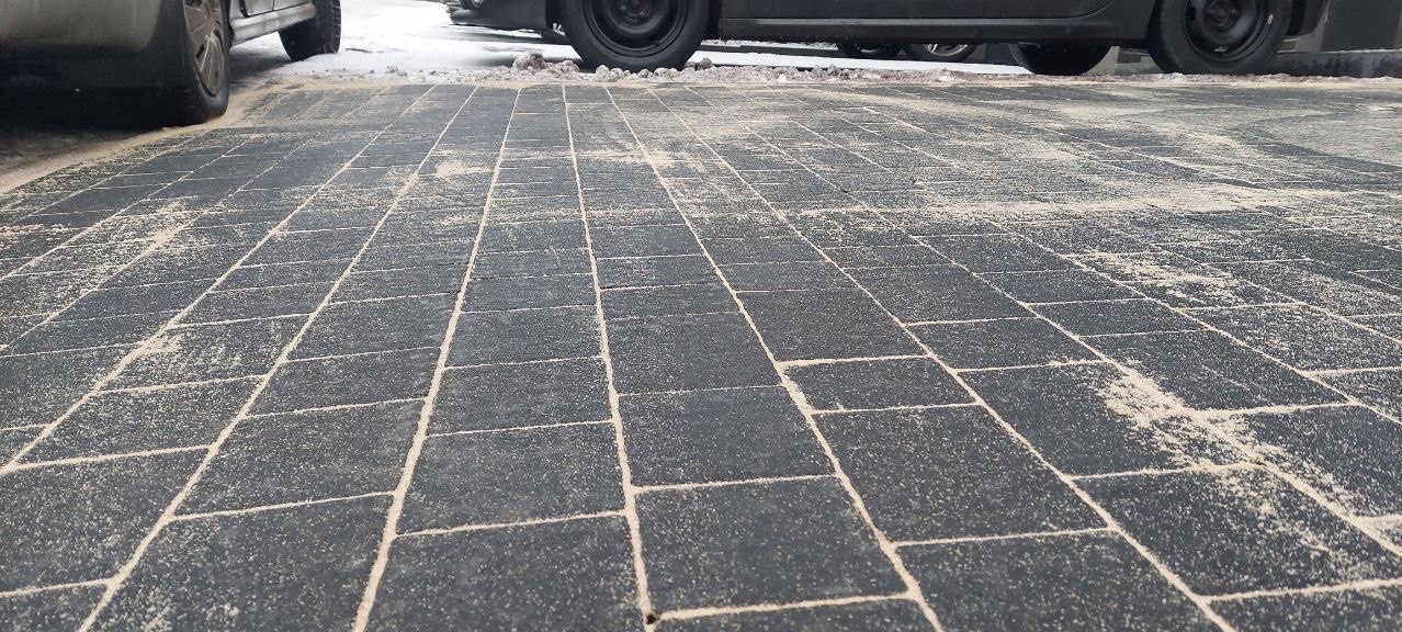 Ułożona, szara kostka brukowa z widocznymi szczelinami wypełnionymi piaskiem, z fragmentami zaparkowanych samochodów w tle.