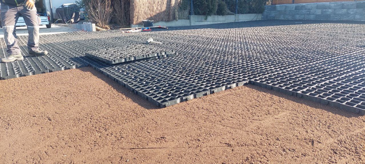 Częściowo ułożona, czarna, plastikowa kratka trawnikowa na podsypce z czerwonego piasku; w tle widoczny pracownik i narzędzia.
