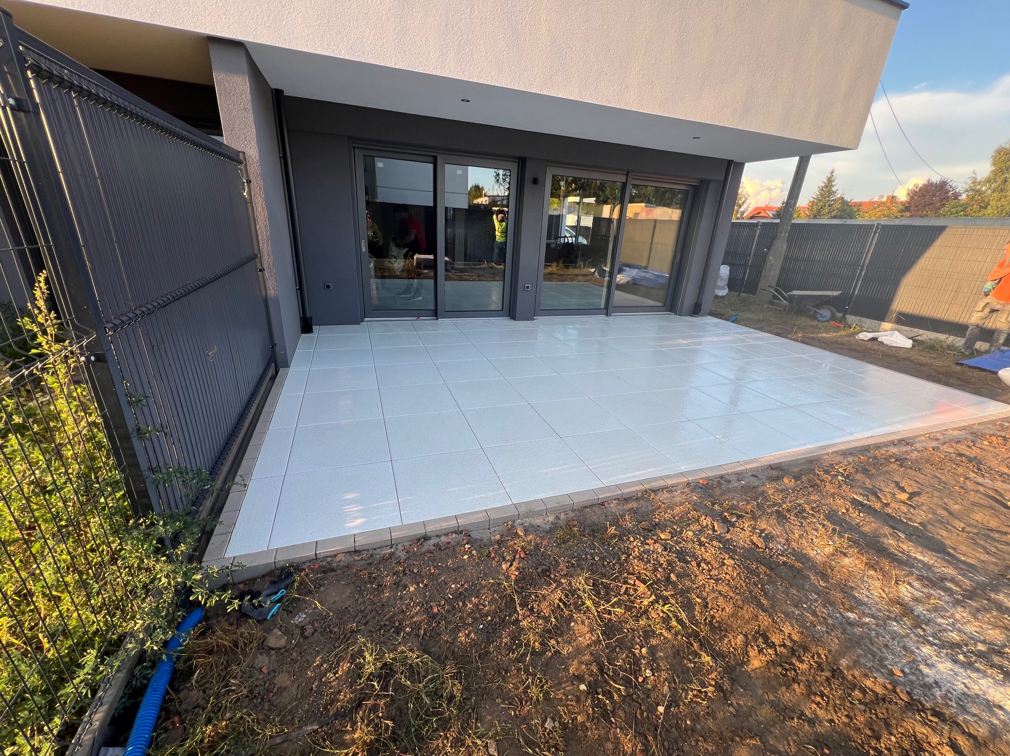 Nowoczesny taras z jasnych, kwadratowych płytek, obramowany kostką. Widok na przesuwne drzwi tarasowe i fragment ogrodu z metalowym ogrodzeniem.