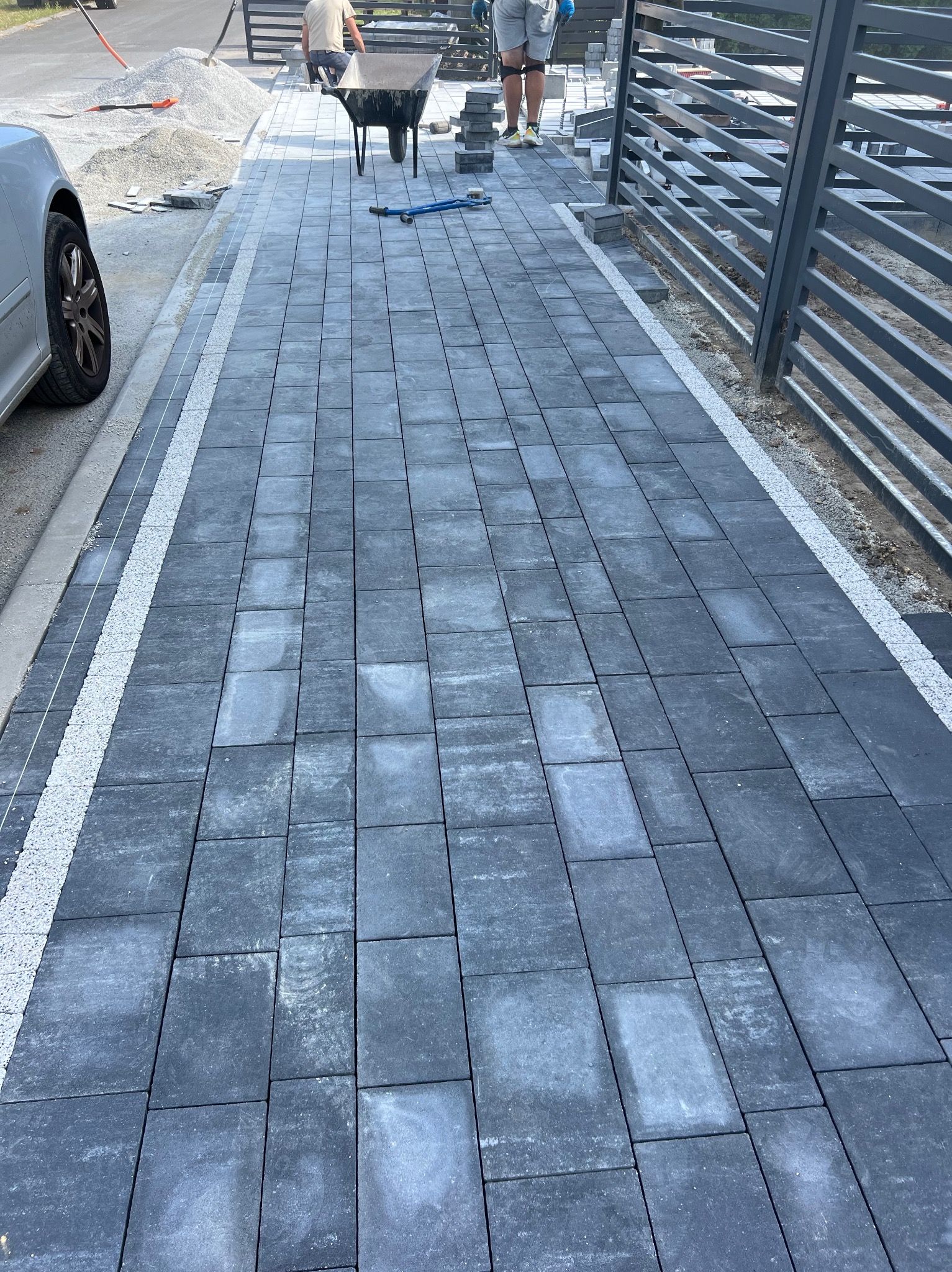 Ułożona, grafitowa kostka brukowa z jasnymi fugami, tworząca ścieżkę wzdłuż metalowego ogrodzenia. W tle widoczny wózek budowlany i materiały do układania.