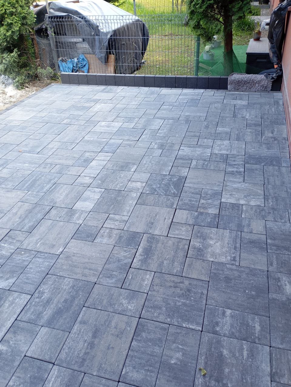 Ułożona szara kostka brukowa na posesji, widok z góry. Różne rozmiary kostek tworzą wzór. W tle siatka ogrodzeniowa i zieleń. Krawężnik z ciemnej kostki.
