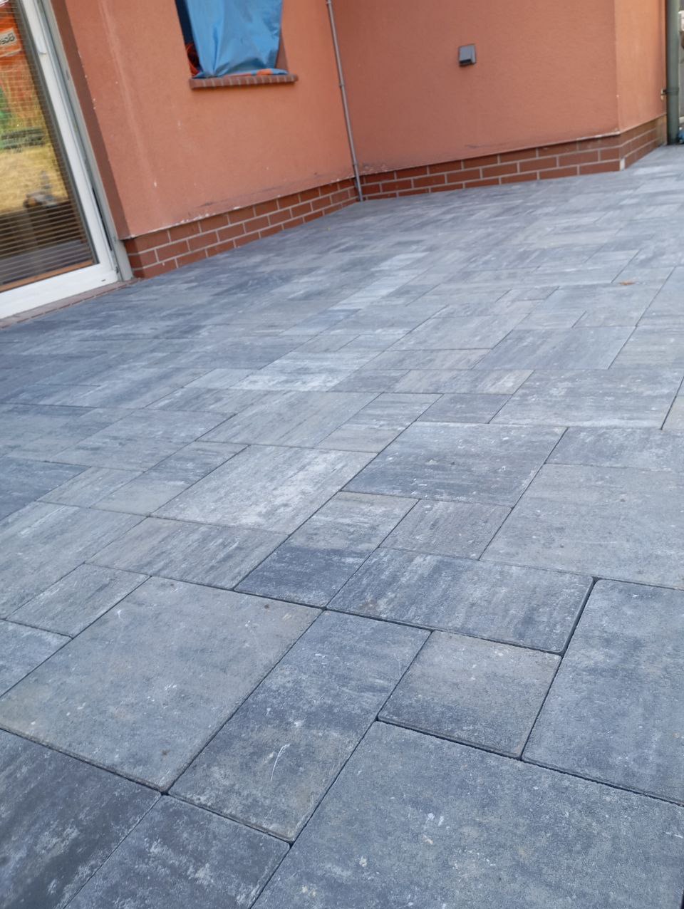 Szara kostka brukowa ułożona na podwórku, widok z góry, z fragmentem elewacji budynku w tle. Regularny wzór, lekko wilgotna powierzchnia.