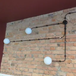 Instalacja elektryczna natynkowa z trzema okrągłymi lampami na ceglanej ścianie z widoczną puszką rozgałęźną i mocowaniami kabli, oświetlenie w stylu retro.
