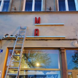BPZ - Metalowa drabina oparta o fasadę budynku z czerwonymi literami 'M' i 'A' oraz oświetleniem, widoczne okna i plakat informujący o otwarciu lokalu.