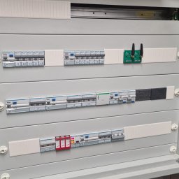 BPZ - Otwarta skrzynka rozdzielcza z bezpiecznikami, wyłącznikami i modułem sterującym z antenami, zamontowana przy ceglanej ścianie.